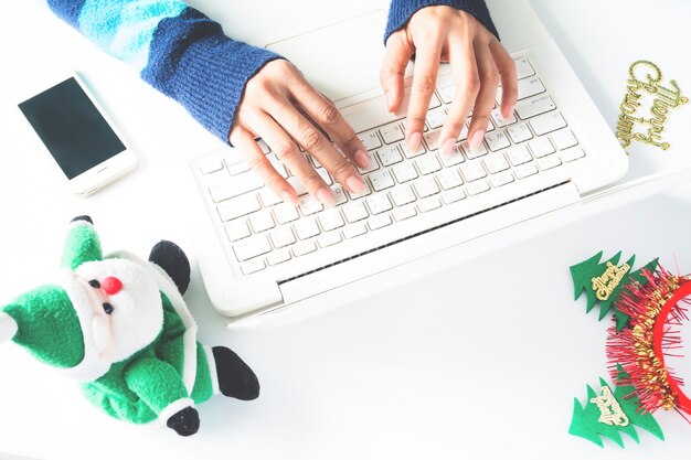 Manos de la mujer escribiendo en el teclado de la computadora portátil, utilizando el teléfono inteligente con la decoración de Navidad, Compras en línea