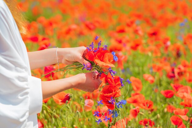 Manos de mujer hermosa joven teje una corona de flores de amapola en el campo en verano