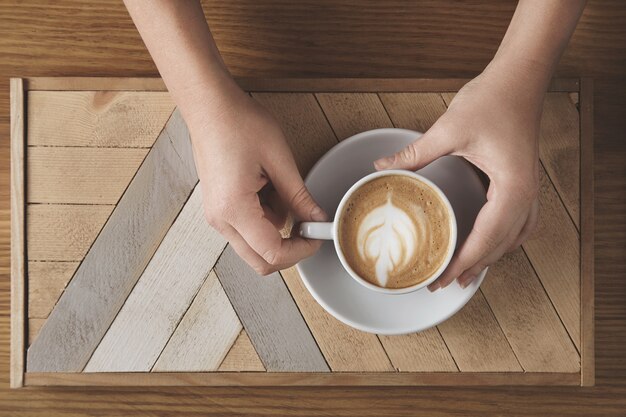 Manos de mujer hermosa sostienen cerámica blanca con capuchino sobre placa de madera y mesa rústica. Espuma de leche en la parte superior en forma de árbol. Vista superior en la cafetería. Concepto de presentación de venta.