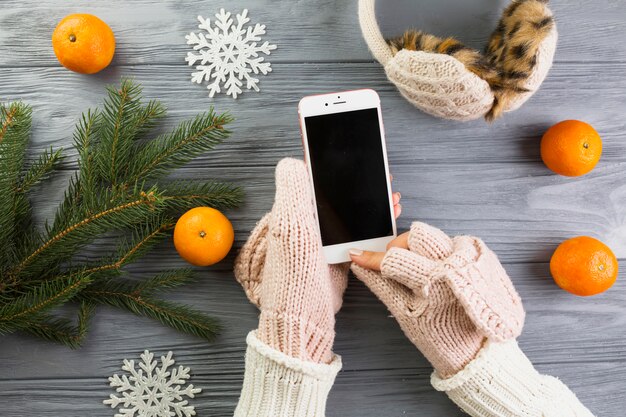 Manos de mujer en mitones con teléfono inteligente cerca de ramas de abeto y copos de nieve de papel