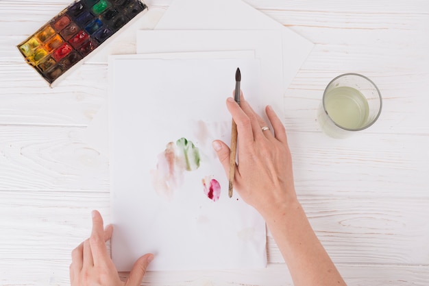 Manos de mujer con pincel cerca de papel con borrones, vidrio y juego de colores de agua