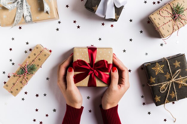 Manos de la mujer que llevan a cabo el arco rojo de la caja de regalo de día de fiesta de la Navidad en la tabla festiva adornada