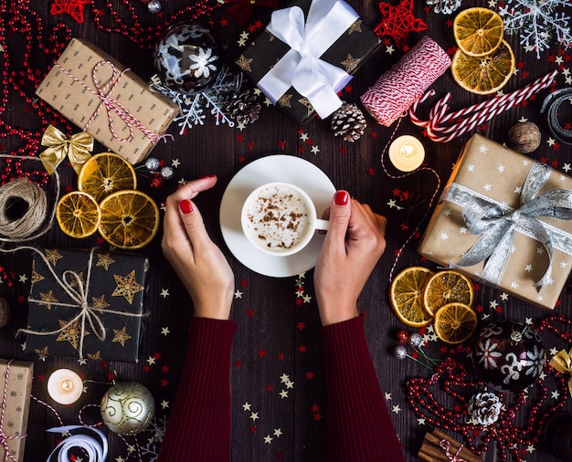 Las manos de la mujer que sostienen la taza de café beben la caja de regalo de vacaciones de la Navidad en la tabla festiva adornada