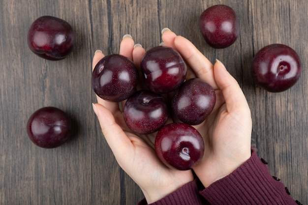 Manos de mujer sosteniendo ciruelas púrpuras saludables en mesa de madera.