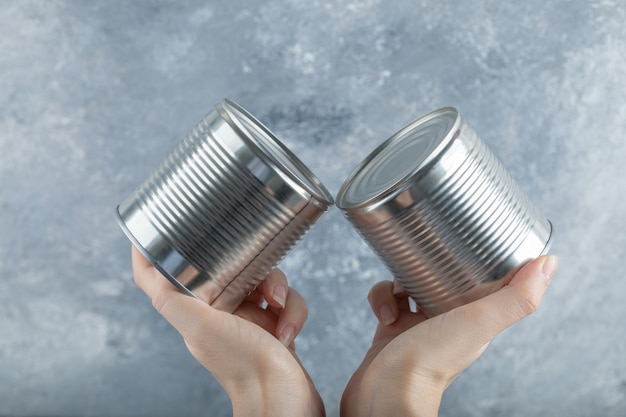 Manos de mujer sosteniendo dos latas metálicas en mármol.