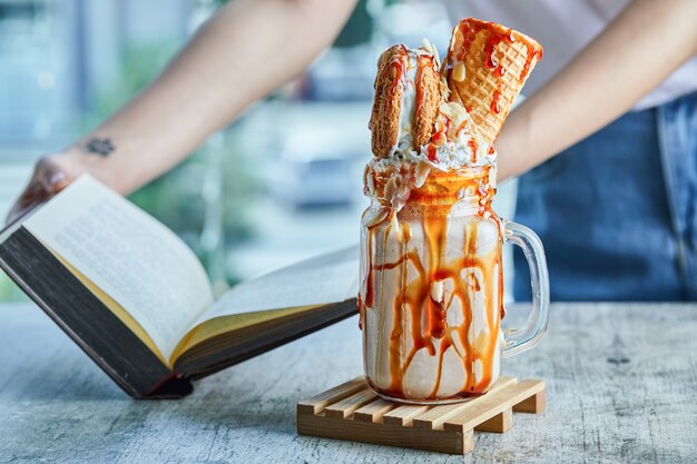 Manos de una mujer con tatuaje sosteniendo un libro y batido de caramelo.