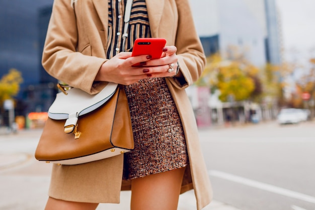 Manos de mujer con teléfono móvil. Chica elegante en abrigo beige charlando. Ciudad moderna.