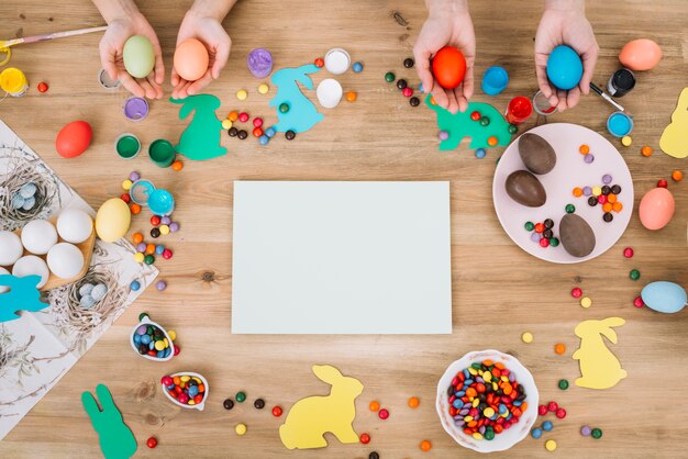 Manos que sostienen los huevos de Pascua con caramelos de colores y papel blanco sobre la mesa de madera