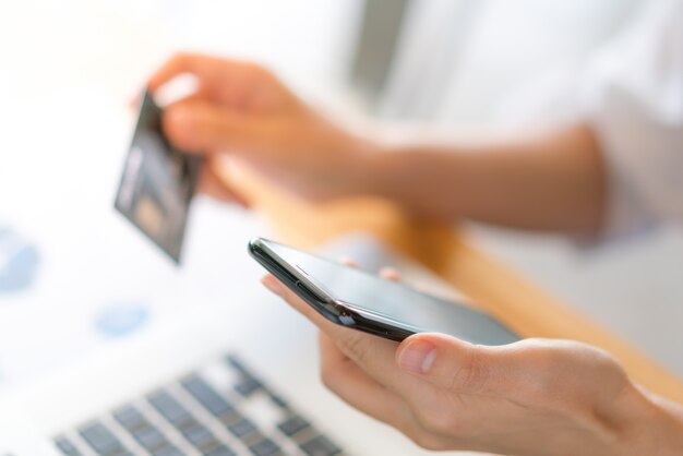 Manos que sostienen una tarjeta de crédito usando la computadora portátil y el teléfono móvil para las compras en línea