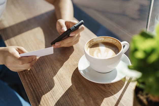 Las manos recortadas de la mujer se apoyan en la mesa de café en el café, se sientan cerca de la ventana, ella sostiene un teléfono inteligente y una tarjeta de crédito o un negocio. La chica envía dinero, ingresa la información de facturación de la cuenta para comprar en línea, beber capuchino.