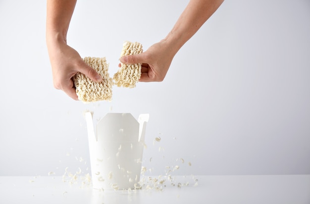 Las manos rompen el paquete prensado de fideos secos sobre la caja de comida para llevar en blanco abierta antes de la preparación. Conjunto de promoción comercial minorista