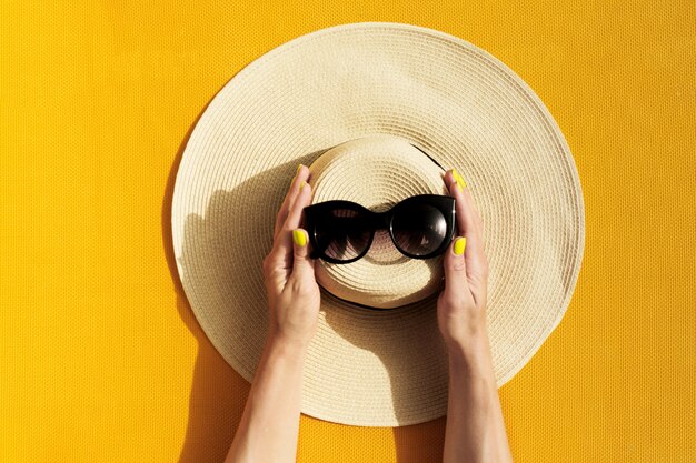 Manos del sombrero de paja de la explotación agrícola de la chica joven y de las gafas de sol en fondo amarillo vibrante.