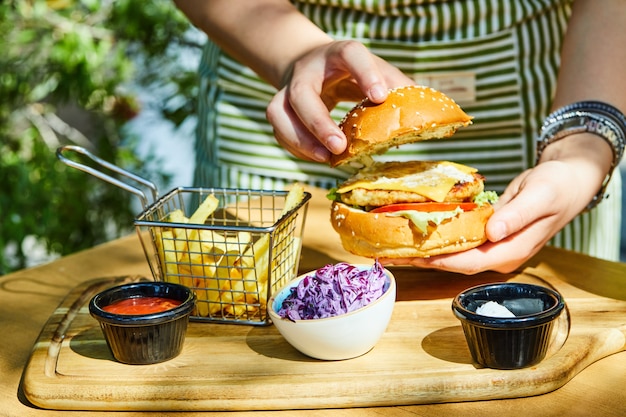 Manos sosteniendo deliciosa hamburguesa fresca con papas fritas y salsa en la mesa de madera.