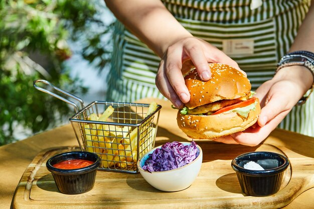 Manos sosteniendo deliciosa hamburguesa fresca con papas fritas y salsa en la mesa de madera.