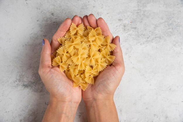 Manos sosteniendo pasta farfalle cruda sobre superficie blanca.