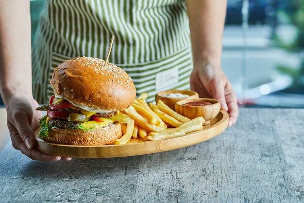 Manos sosteniendo un plato de madera con hamburguesa y patatas fritas con salsa de tomate y mayonesa.