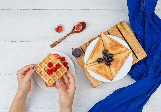 Manos sostienen waffle con bayas y frutas en un plato blanco sobre una superficie blanca