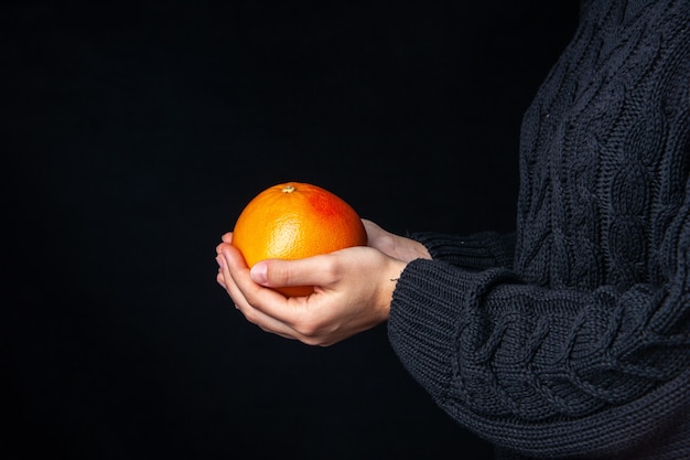 Manos de vista frontal sosteniendo naranja fresca sobre superficie oscura