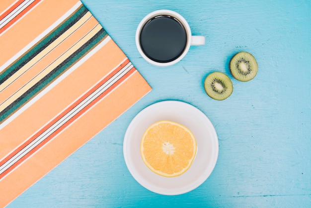 Foto gratuita manteles; taza de café; kiwi a la mitad y fruta naranja sobre fondo azul