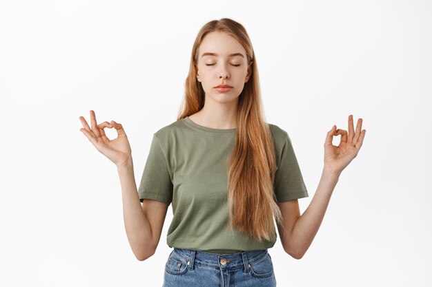 Mantén la calma. Mujer joven relajada meditando, cerrar los ojos y tomarse de las manos hacia los lados en el signo zen mudra, practicar yoga, respirar y descansar la mente, de pie sobre fondo blanco