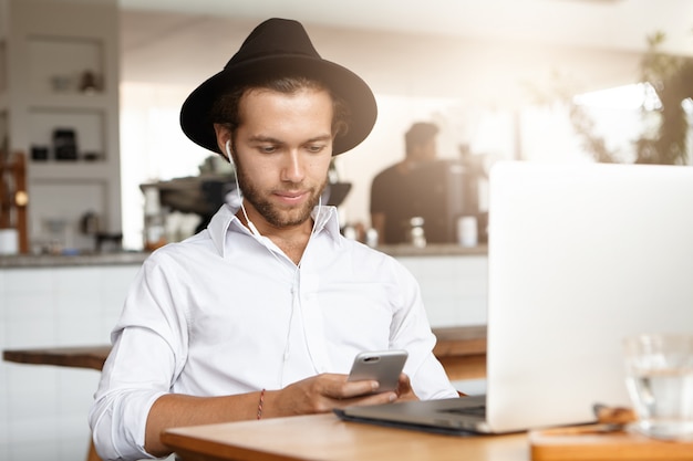 Mantenerse conectado. Apuesto joven con smartphone, sentado en el café frente a la computadora portátil en auriculares mientras espera su almuerzo