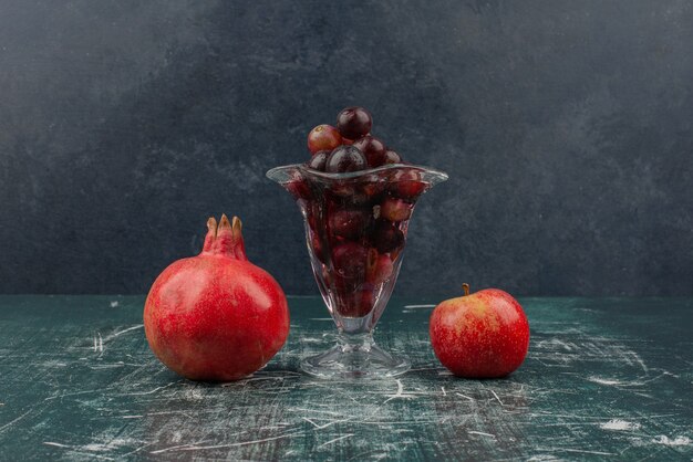 Manzana, granada y copa de uvas negras sobre mesa de mármol.