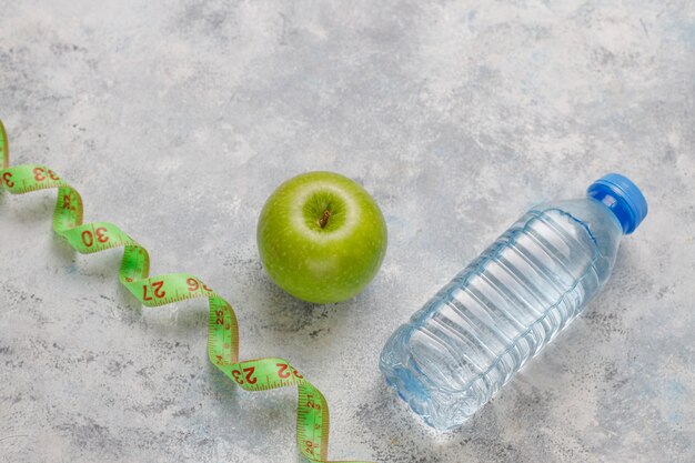 Manzana verde fresca, cinta métrica y botella de agua dulce sobre hormigón gris