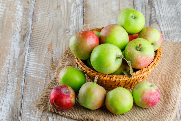 Manzanas en una canasta de mimbre sobre madera y pedazo de saco. vista de ángulo alto.