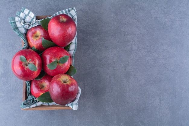 Manzanas con hojas sobre una toalla en una caja sobre una mesa de mármol.