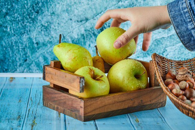Manzanas y peras en canasta de madera y cuenco de avellanas sobre superficie azul.