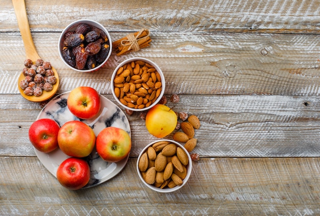 Manzanas en un plato con palitos de canela, dátiles, almendras peladas y sin pelar en tazones, nueces en la cuchara de madera vista superior sobre un fondo de madera