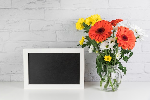 Foto gratuita manzanilla amarilla y blanca con flores rojas de gerbera en el jarrón cerca del marco negro en el escritorio