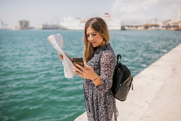 Mapa femenino de la tenencia del turista que mira el teléfono móvil que se coloca cerca del mar