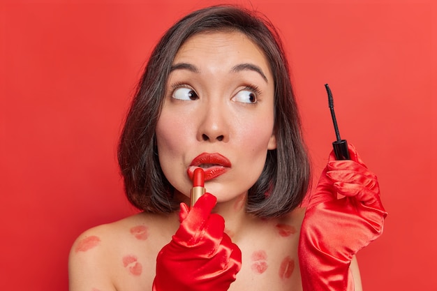 Maquillaje de belleza. La dama asiática se aplica lápiz labial y rímel usa cosméticos decorativos para una apariencia fabulosa usa guantes rojos posa desnuda contra la pared brillante del estudio se prepara para una ocasión especial