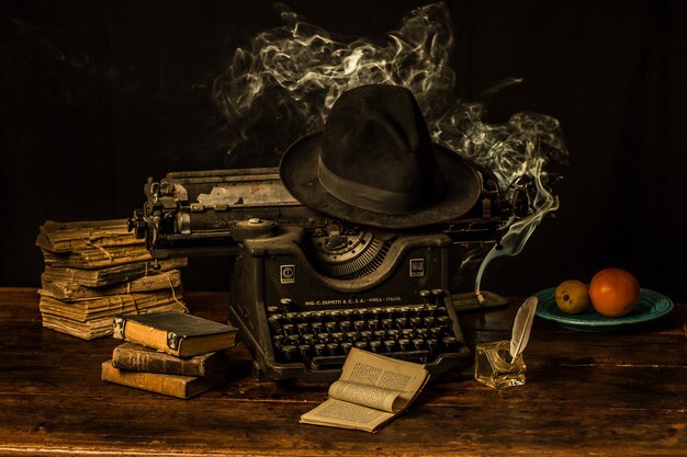 Una máquina de escribir, un sombrero fedora y libros antiguos sobre una mesa de madera