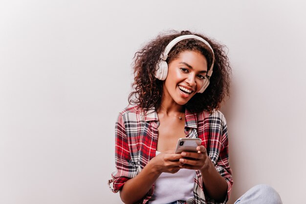 Maravillosa chica africana con mensaje de texto de sonrisa linda. Atractiva mujer feliz en auriculares blancos posando en beige