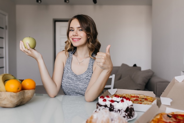 Maravillosa chica blanca disfrutando de su dieta con frutas frescas. Retrato interior de mujer impresionante elige entre manzana y pastel.