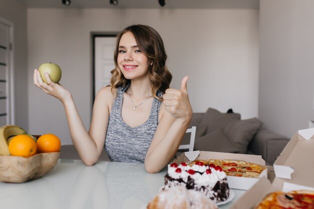 Maravillosa chica blanca disfrutando de su dieta con frutas frescas. Retrato interior de mujer impresionante elige entre manzana y pastel.