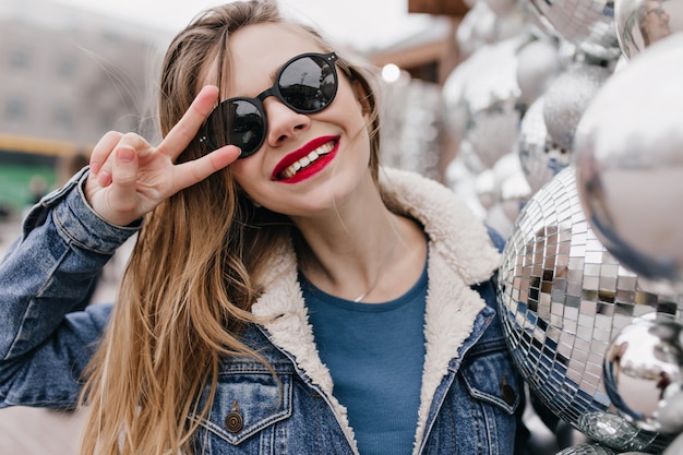 Maravillosa chica blanca con gafas de sol posando con el signo de la paz en un frío día de primavera. Tiro al aire libre de modelo de mujer riendo en chaqueta de mezclilla divirtiéndose en la mañana.