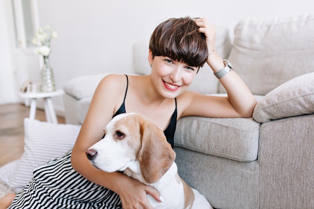 Maravillosa chica bronceada apoyando la cabeza con la mano posando en casa después de un divertido juego con perro beagle