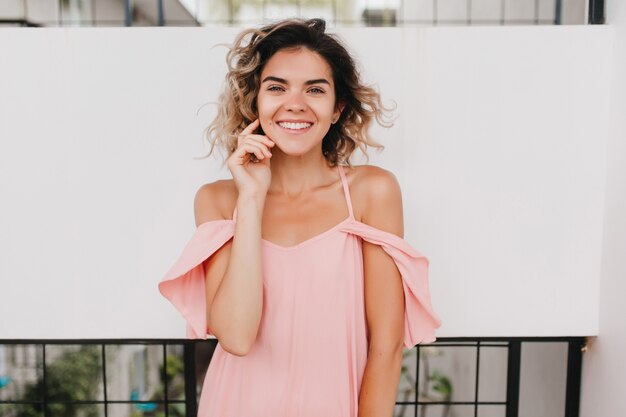 Maravillosa chica bronceada en traje de verano rosa tocando su rostro. Retrato de mujer joven delgada riendo con piel bronceada.