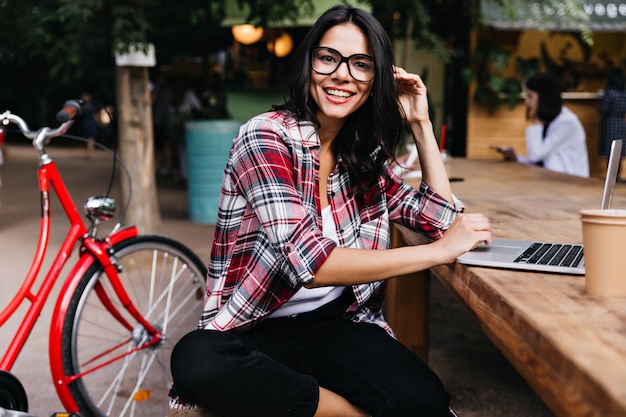 Maravillosa chica de buen humor sentada en la ciudad con portátil y sonriendo. Retrato al aire libre de una atractiva dama morena con gafas posando al lado de la bicicleta.