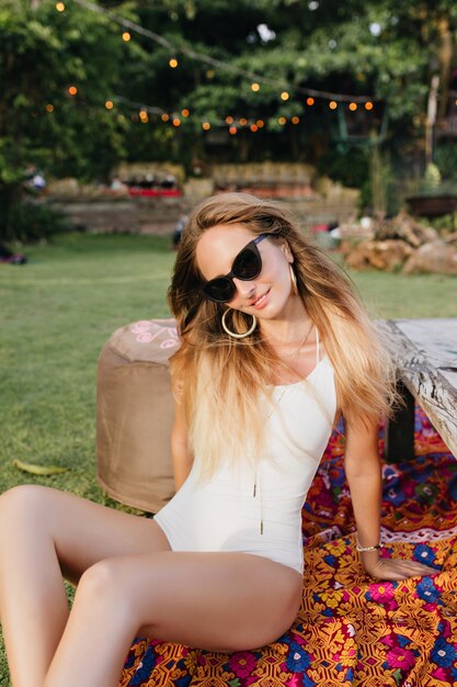 Maravillosa chica caucásica posando con sonrisa interesada mientras toma el sol en la hierba. Linda mujer rubia en traje de baño blanco tirado en el suelo en el parque.