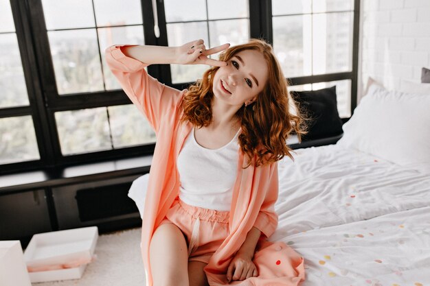 Maravillosa chica en pijama lindo sonriendo en su piso. Filmación en interiores de fascinante mujer jengibre divirtiéndose en la mañana.