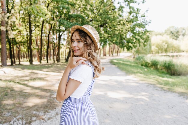 Maravillosa chica rubia caminando en el parque de verano y sonriendo. Foto al aire libre de la adorable dama con sombrero de paja posando con placer en la naturaleza.