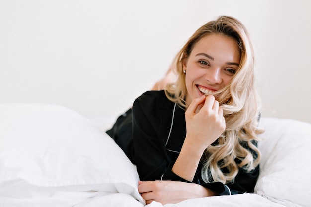 Maravillosa chica rubia con estilo en traje de dormir negro acostado en la cama y sonriendo