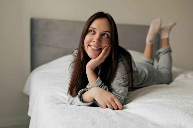 Maravillosa chica soñadora con cabello oscuro está acostada en la cama con una sonrisa feliz en un estado de ánimo romántico. La dama encantadora se despertó por la mañana y planeó su día.