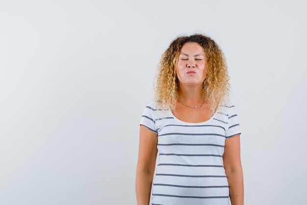 Maravillosa dama haciendo pucheros con los labios mientras cierra los ojos en una camiseta a rayas y luce positiva, vista frontal.