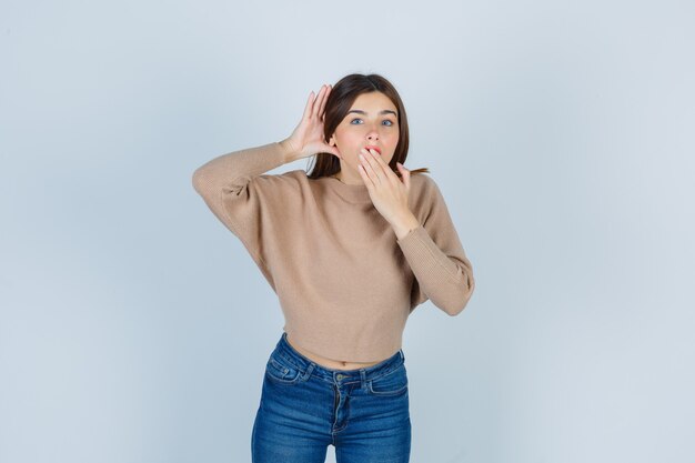 Maravillosa dama en suéter, jeans manteniendo una mano detrás de la oreja y la otra en la boca y mirando sorprendida, vista frontal.