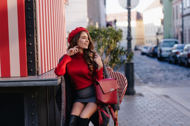 Maravillosa dama viste minifalda sentada en un café al aire libre con mochila roja y mirando a su alrededor